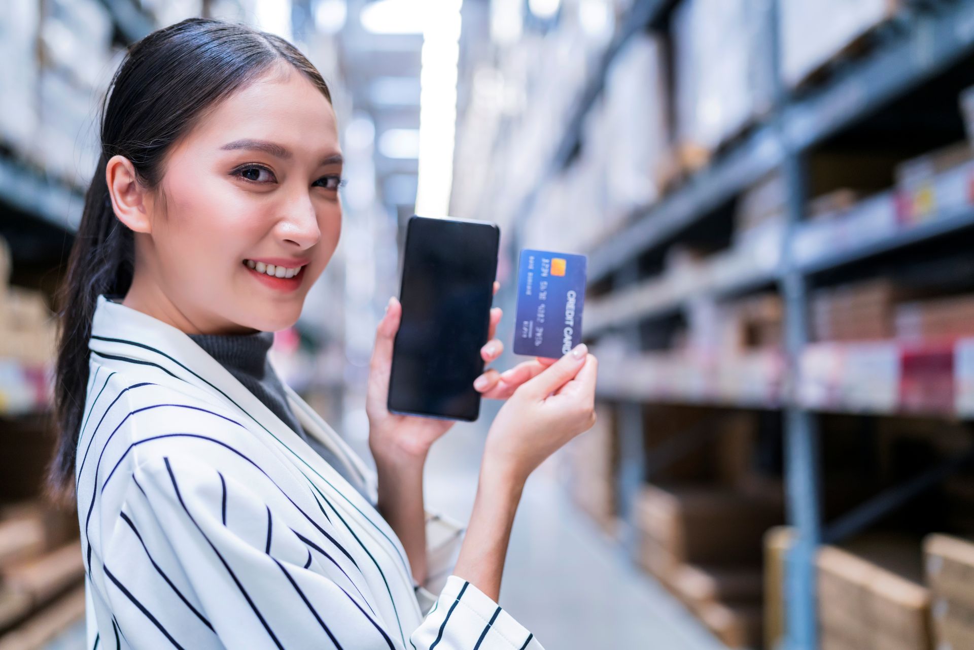 信用卡換現金示意圖：女性在倉儲走道手持智慧手機與信用卡，呈現線上刷卡交易與資金周轉情境，適用於信用卡換現金、費用試算與風控避雷主題